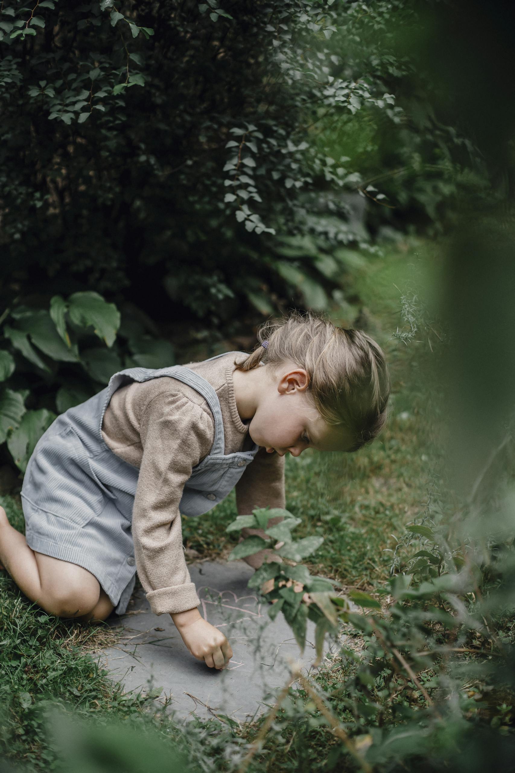 A little girl attentively drawing with chalk on a stone path surrounded by greenery.