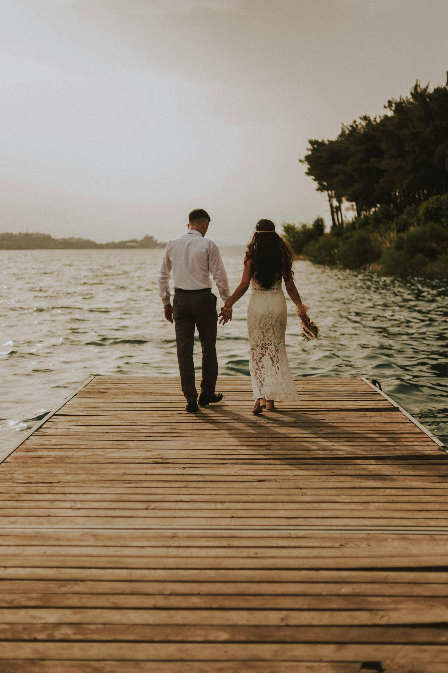 Newlyweds holding hands and walking on a wooden dock by the lake during sunset in Adana, Türkiye.