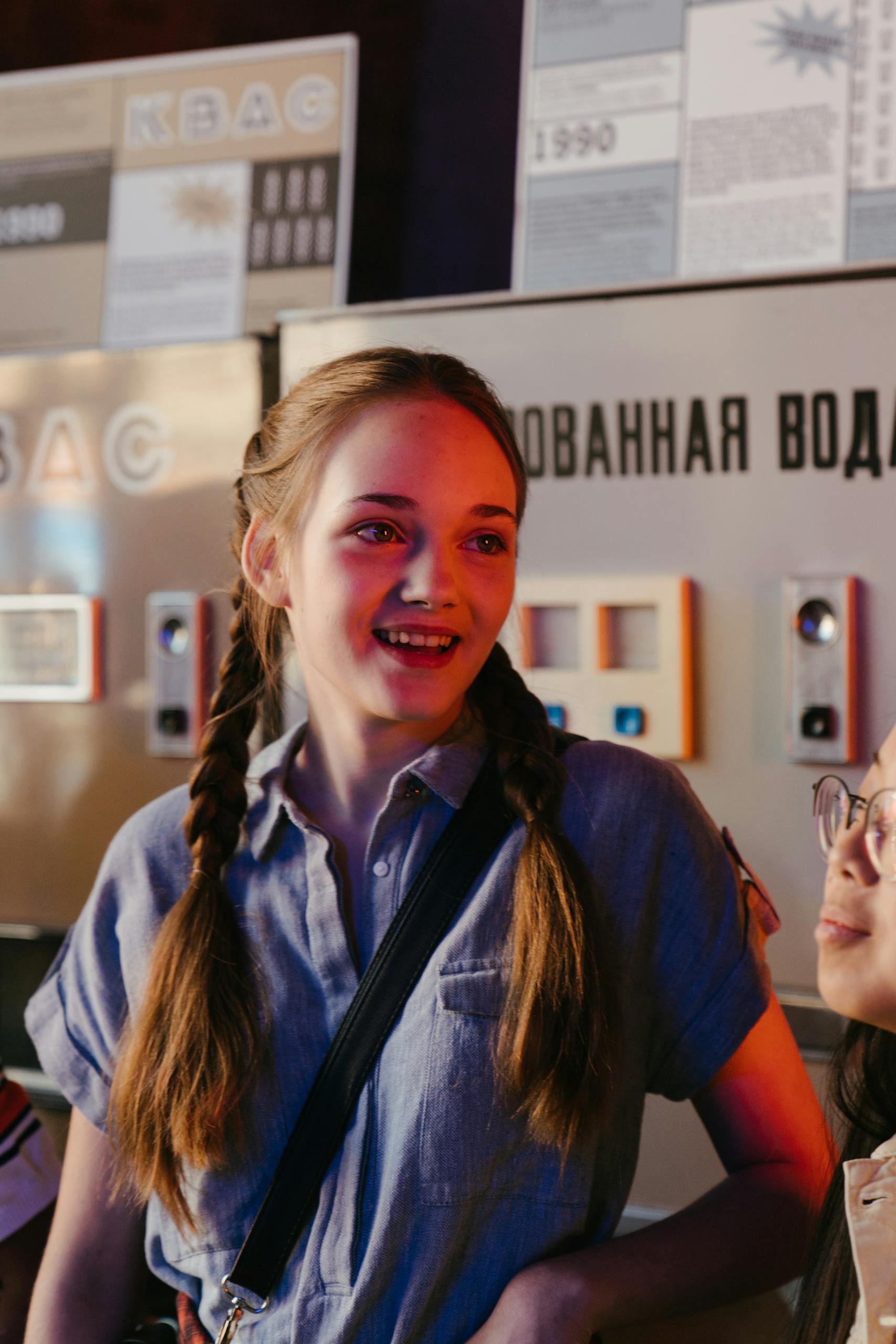 Smiling teenage girl in braids stands near vintage vending machine under warm lighting.