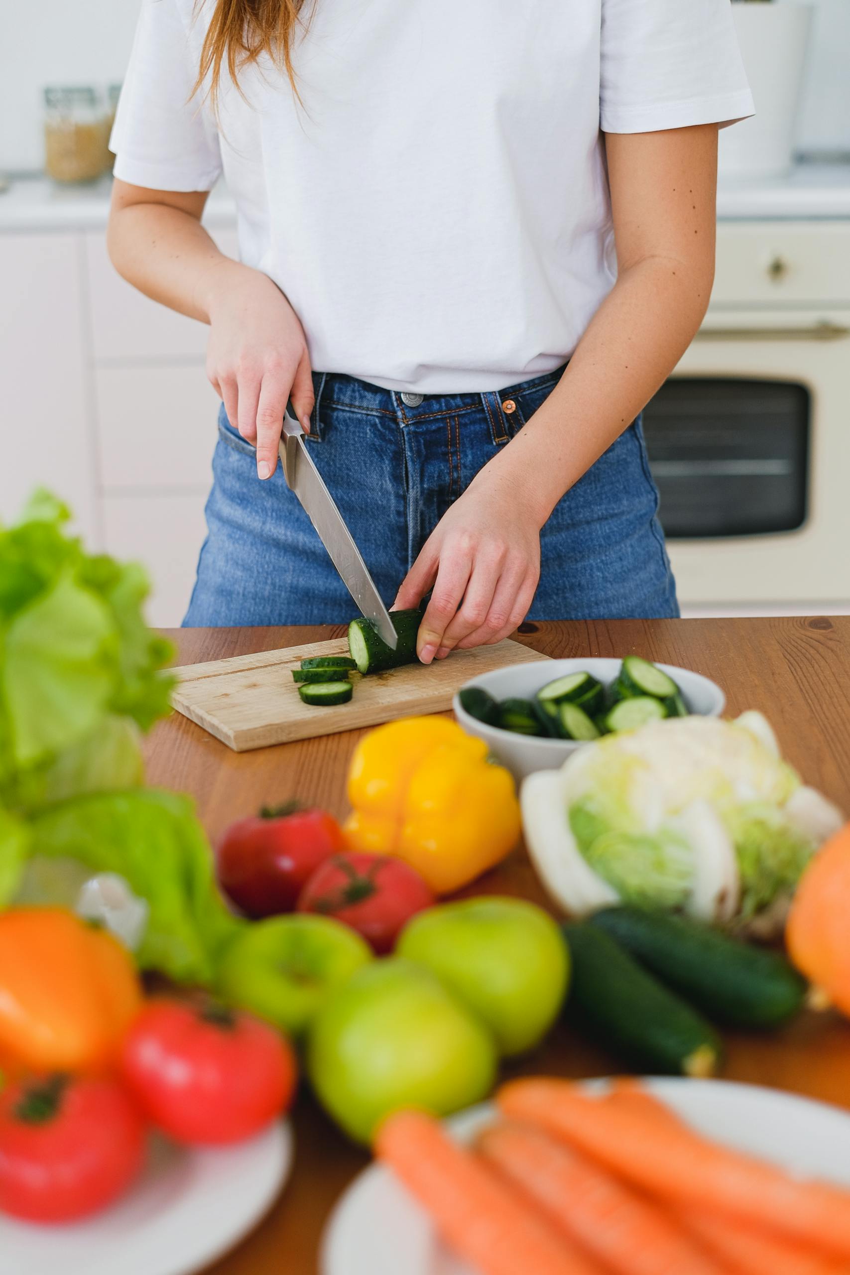 Woman slicing cucumbers in a kitchen surrounded by fresh vegetables, exemplifying healthy eating.